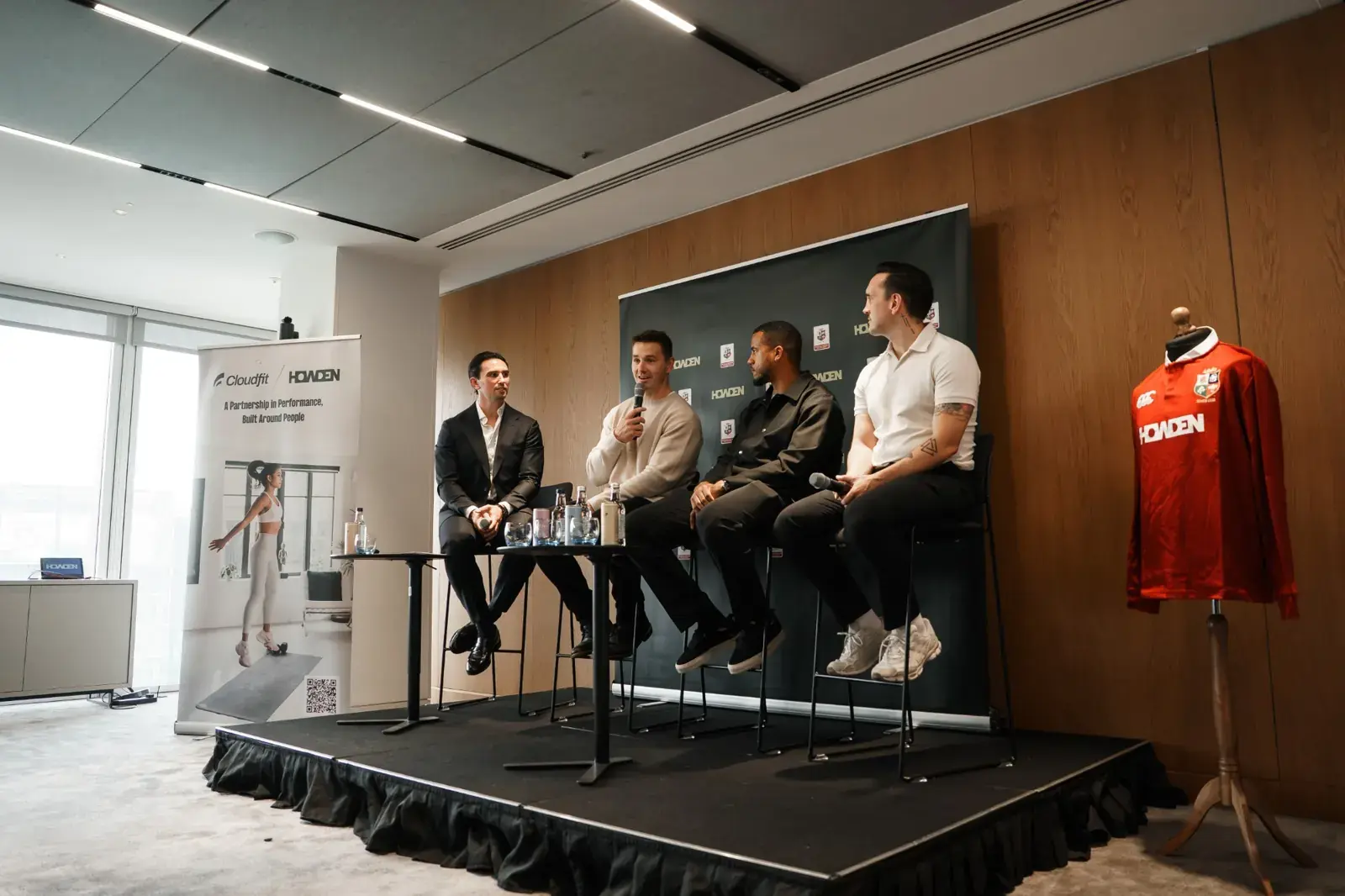 A panel on stage at the CloudFit and Howden event, beside a CloudFit and Howden banner and a British and Irish Lions shirt